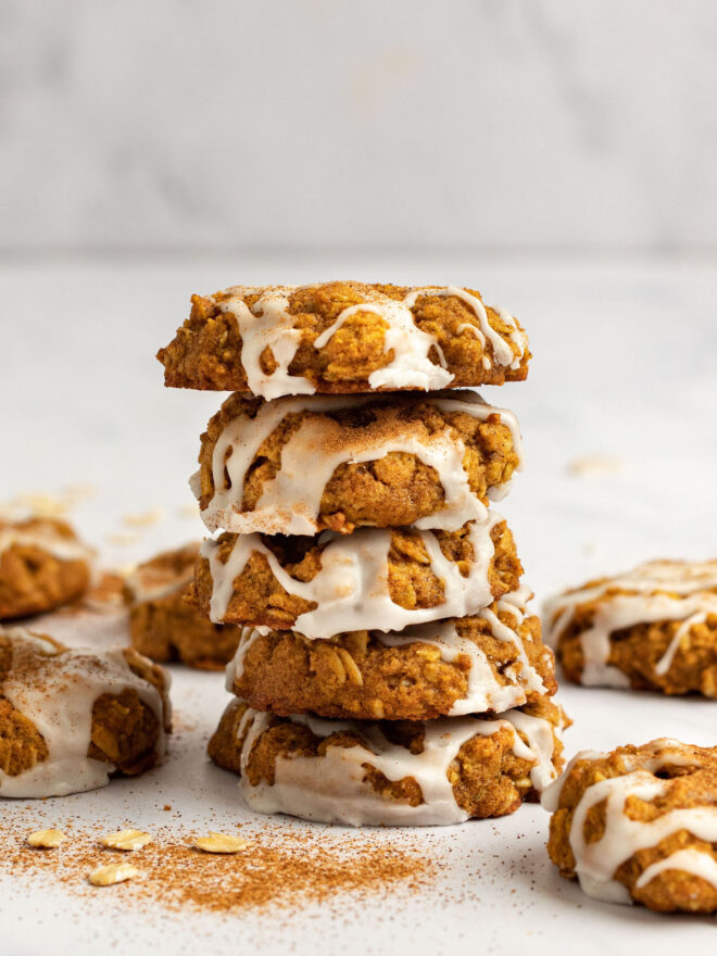 This is a vertical image looking at a stack of five pumpkin cookies from the side. The cookies are each drizzled with a white, hardened icing and sprinkled with a pumpkin spice. The stack sits on a white surface with more cookies on the counter around the stack. Blurred in the background is a white marble backsplash.