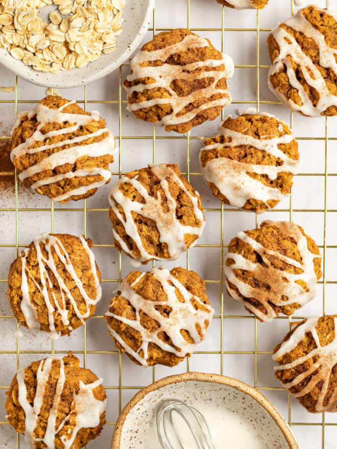 This is an overhead vertical image of pumpkin oat cookies drizzled with a white icing and sprinkled with pumpkin spice. The cookies sit on a gold cooling rack. A small white speckled bowl with oats is to the top left corner of the image and a small white speckled bowl with a brown rim is in the bottom center of the image with white icing and small whisk in it. The cooling rack sits on a white surface.