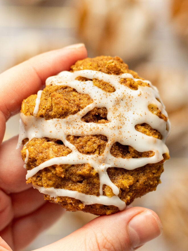 This is a vertical image focusing closeup on a left hand holding a pumpkin oatmeal cookie. The cookie is drizzled with white icing and a pumpkin spice. More cookies are blurred in the background.