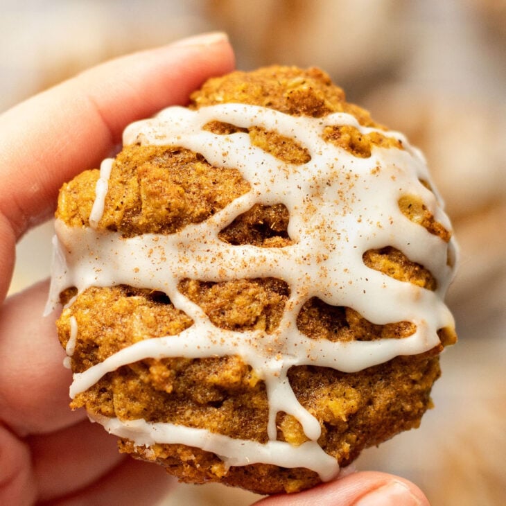 This is a vertical image focusing closeup on a left hand holding a pumpkin oatmeal cookie. The cookie is drizzled with white icing and a pumpkin spice. More cookies are blurred in the background.