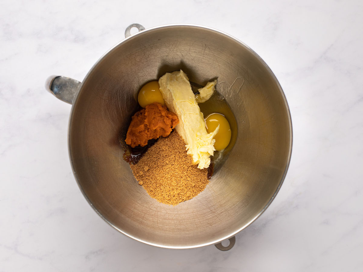This is a horizontal overhead image looking into a silver mixing bowl with butter, eggs, pumpkin puree, and coconut sugar. The mixing bowl sits on a white marble surface.