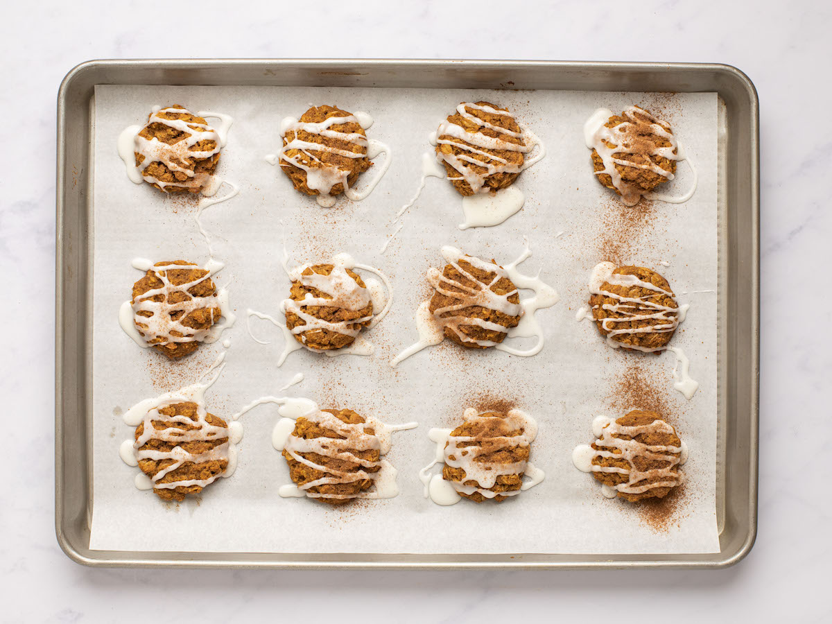 This is an overhead horizontal image looking at a silver rimmed baking sheet lined with parchment paper. On the parchment paper are 12 baked oatmeal cookies drizzled with a white icing. The baking sheet sits on a white marble surface.