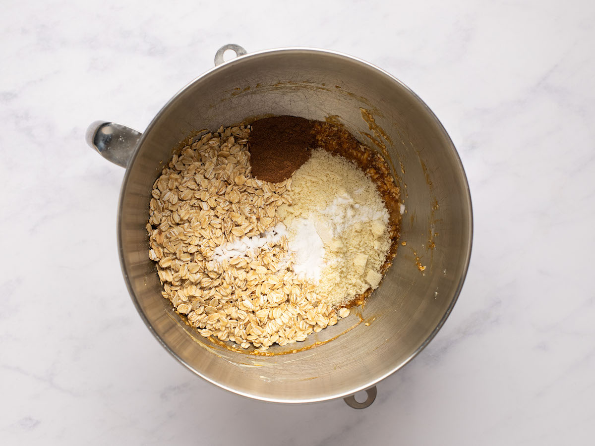 This is a horizontal overhead image looking into a silver mixing bowl with almond flour, oats, spice, and baking soda on top of wet ingredients. The mixing bowl sits on a white marble surface.