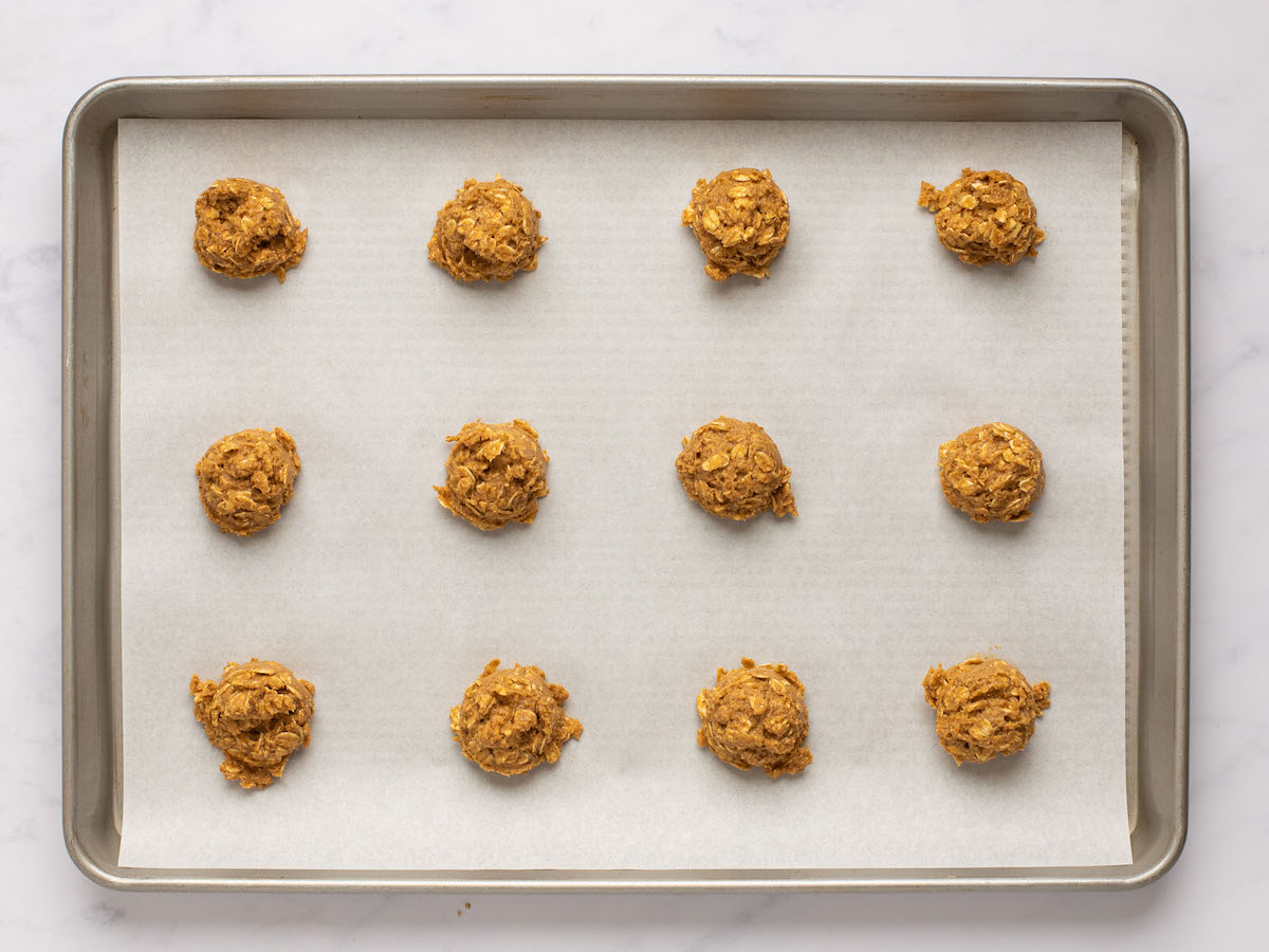 This is an overhead horizontal image looking at a silver rimmed baking sheet lined with parchment paper. On the parchment paper are 12 oatmeal cookie dough balls spaced apart. The baking sheet sits on a white marble surface.