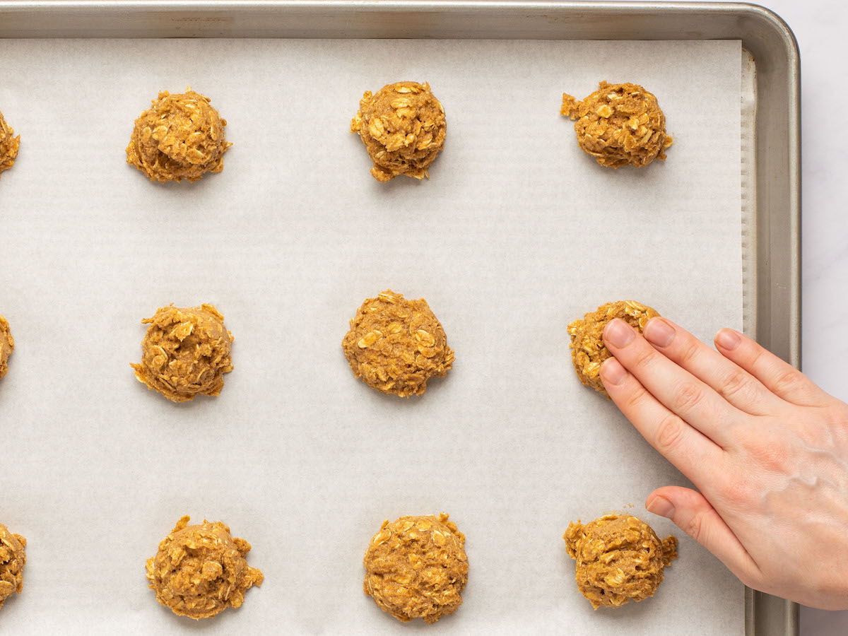 This is an overhead horizontal image looking at a silver rimmed baking sheet lined with parchment paper. On the parchment paper are 12 oatmeal cookie dough balls spaced apart. The baking sheet sits on a white marble surface. A right hand is coming from the bottom right corner of the image pressing the top of the middle cookie dough ball to flatten it.