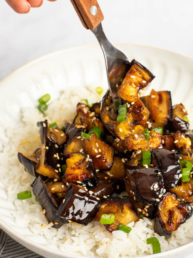 This is a vertical image looking at a shallow white bowl from the side. In the bowl is white rice and fried golden brown eggplant pieces. The eggplant are sprinkled with sesame seeds and chopped scallions. A fork is coming from the top of the image and piercing some pieces of eggplant.