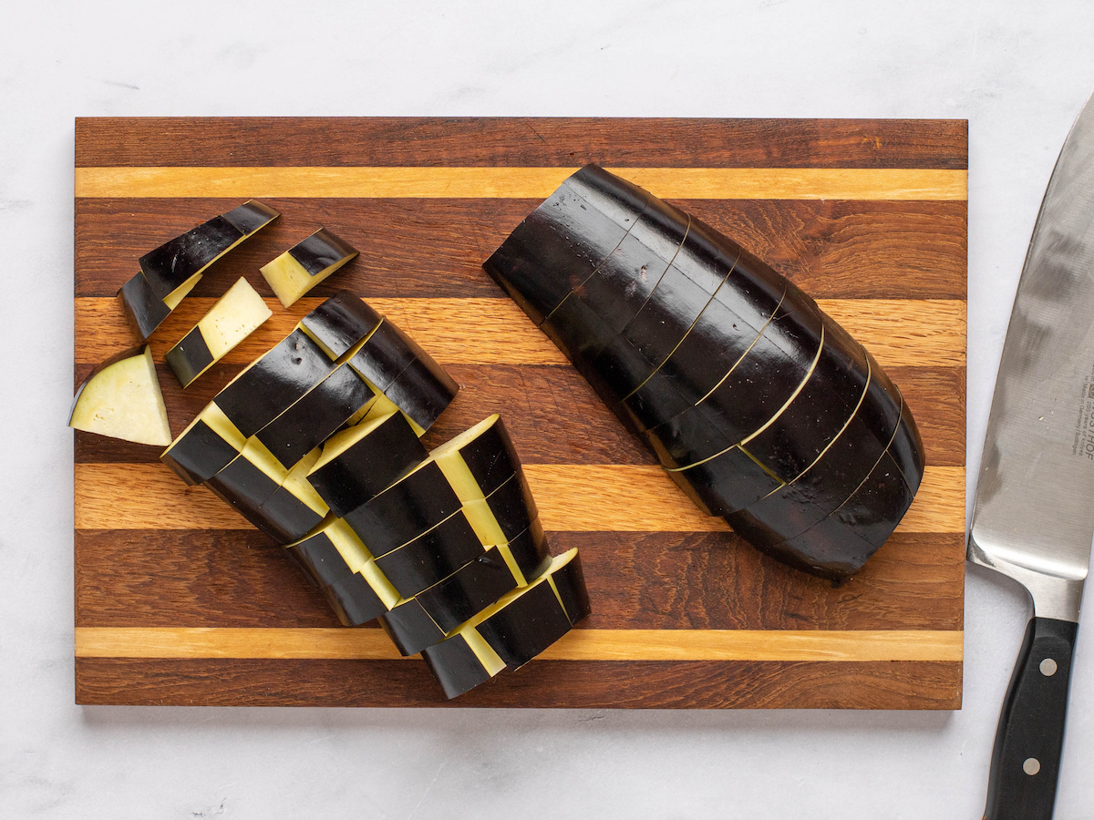This is an overhead horizontal image of a raw eggplant cut into cubes on a wood striped cutting board. The cutting board sits on a light marble surface. A sharp knife is to right right side of the image.