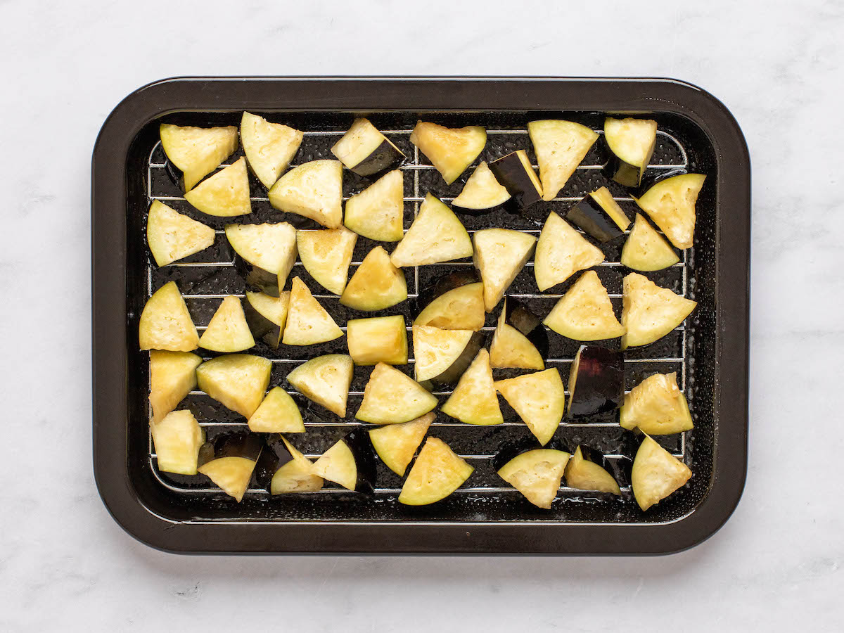 This is an overhead horizontal image of triangles of raw eggplant spread out in an even layer on an air fryer try. The tray sits on a white marble surface. The eggplant pieces appear to have been sprayed with oil.