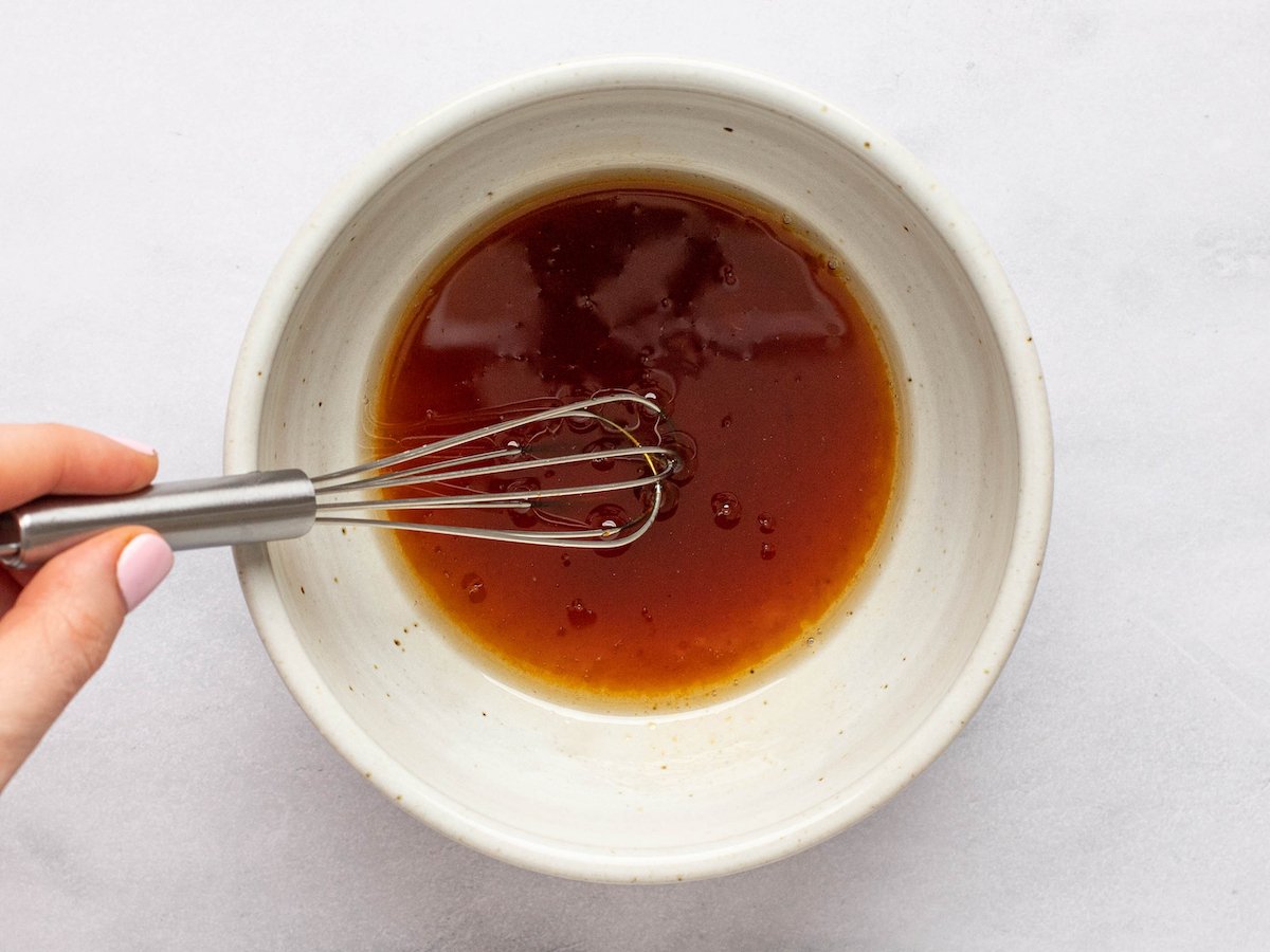 This is an overhead horizontal image of a small white bowl on a white counter. In the bowl is a soy mixture. A hand is coming in the image from the left side and holding a small whisk. The whisk is dipping into the sauce.