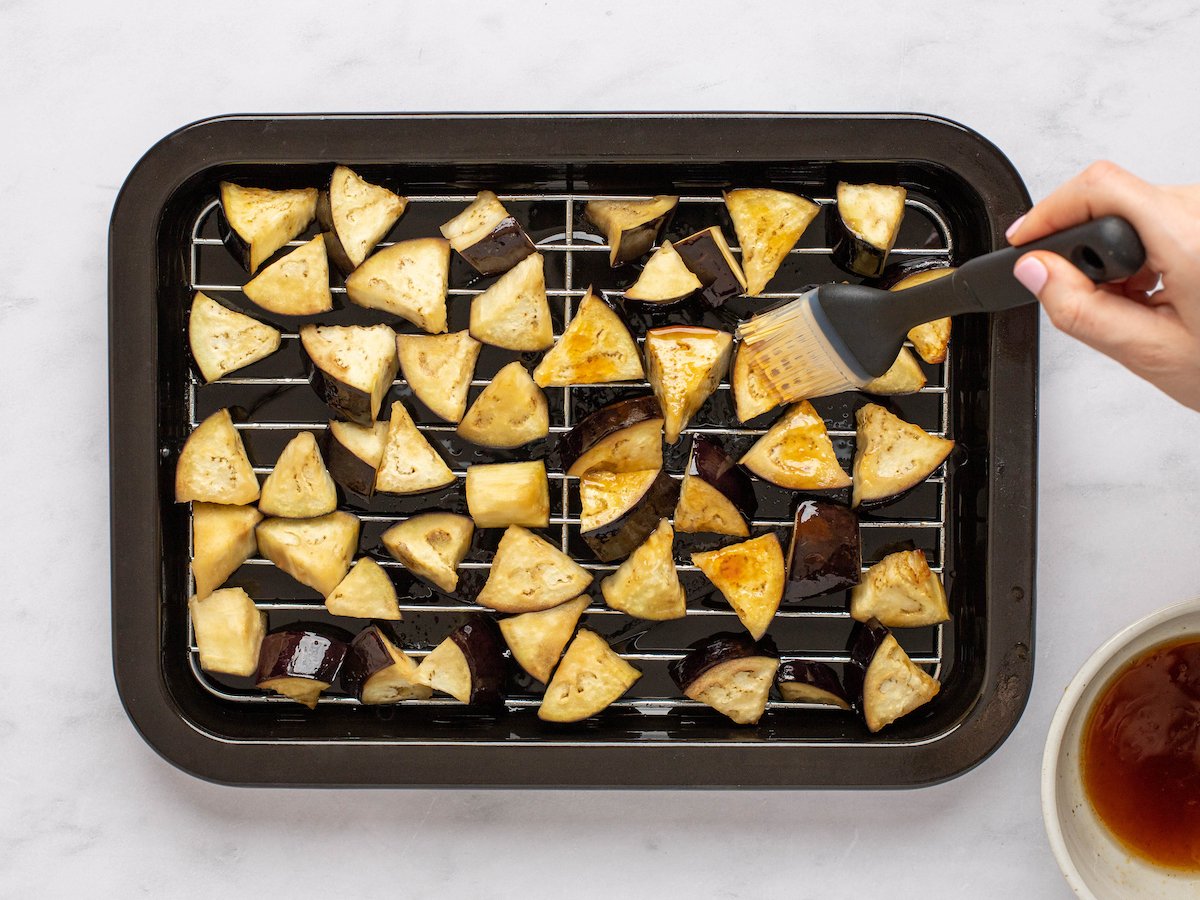 This is an overhead horizontal image of triangles of eggplant spread out in an even layer on an air fryer try. The tray sits on a white marble surface. A hand is coming in the right side of the image and holding a silicone pastry brush. The brush is applying a light brown sauce to the eggplant pieces.