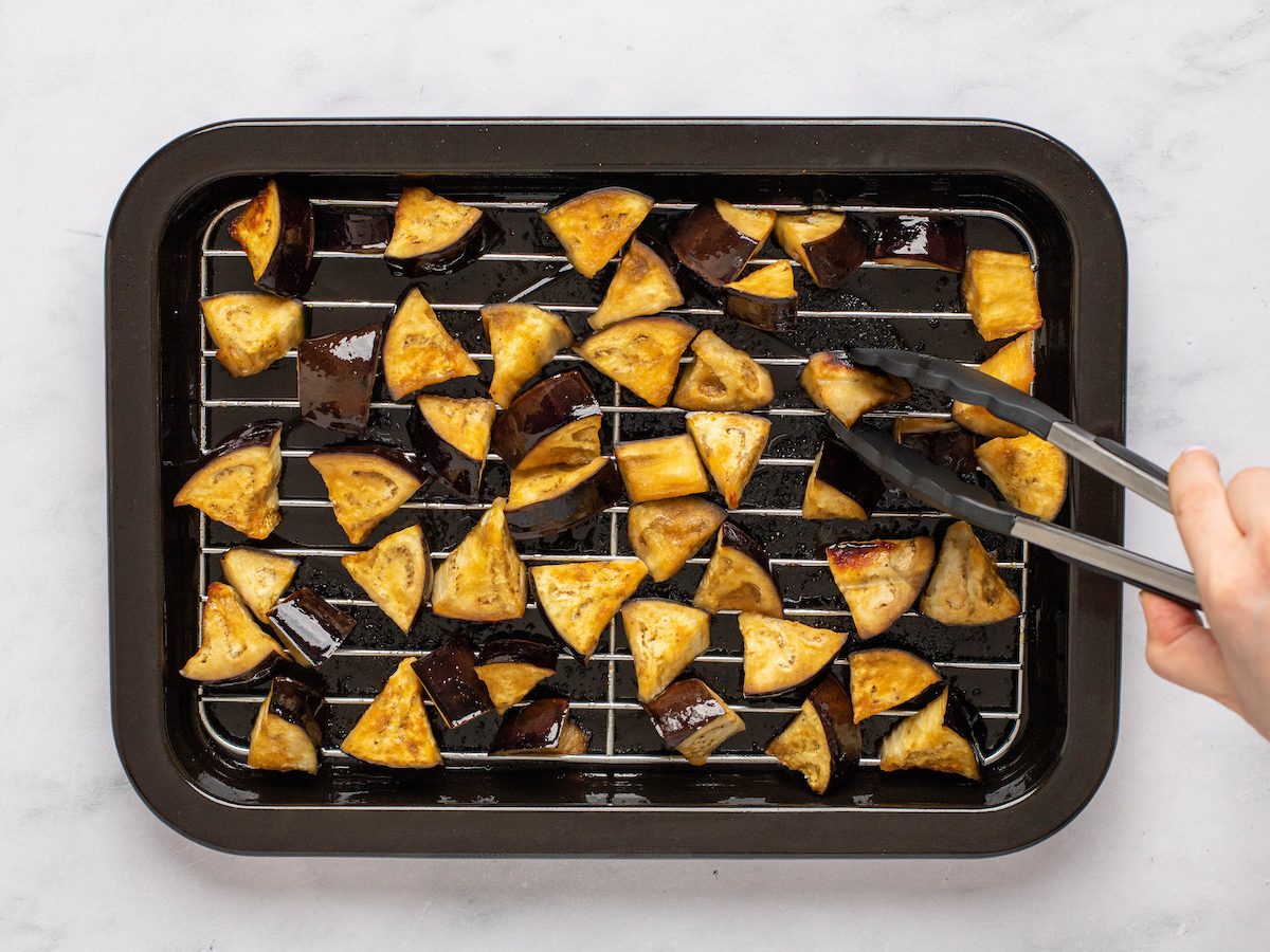 This is an overhead horizontal image of triangles of raw eggplant spread out in an even layer on an air fryer try. The tray sits on a white marble surface. A hand is coming in from the right side of the image and holding tongs that are picking up a piece of the eggplant to be flipped.