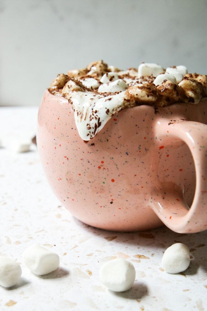 This is a vertical image looking at a speckled pink mug from the side. The mug is overflowing with marshmallow fluff and mini marshmallows which are topped with shaved chocolate. A drop of marshmallow fluff is dripping down the side of the mug. The mug sits on a white and tan terrazzo surface with a white marble surface in the background. Scattered around the mug are mini marshmallows.