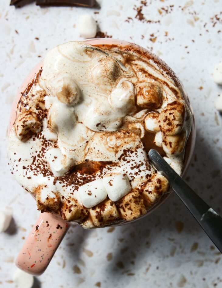 This is an overhead vertical image of a pink speckled mug filled with a chocolate, marshmallow and fluff mixture. The mug is sitting on a white and tan terrazzo surface. The mug is topped with shaved chocolate and more pieces of chocolate bar and mini marshmallows are on the counter around the mug. A silver spoon is in the mug with the handle of the spoon pointing to the right bottom corner of the image.