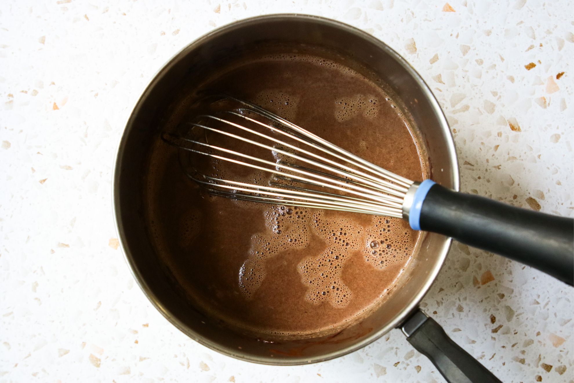 This is an overhead horizontal image of a stainless steel saucepan with a chocolate liquid in it. The pan sits on a white and tan terrazzo surface. A silver whisk with a black rubber handle is in the pan with the handle leaning against the side of the pan. The handle is pointing to the right side of the image.