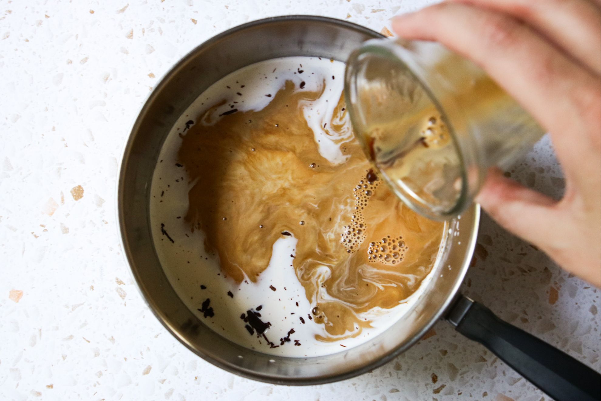 This is an overhead horizontal image of a stainless steel saucepan with milk and espresso being poured into it. A hand is coming in from the right side of the image tilting a small glass cup of espresso and pouring it into the saucepan. The pan sits on a white and tan terrazzo surface.