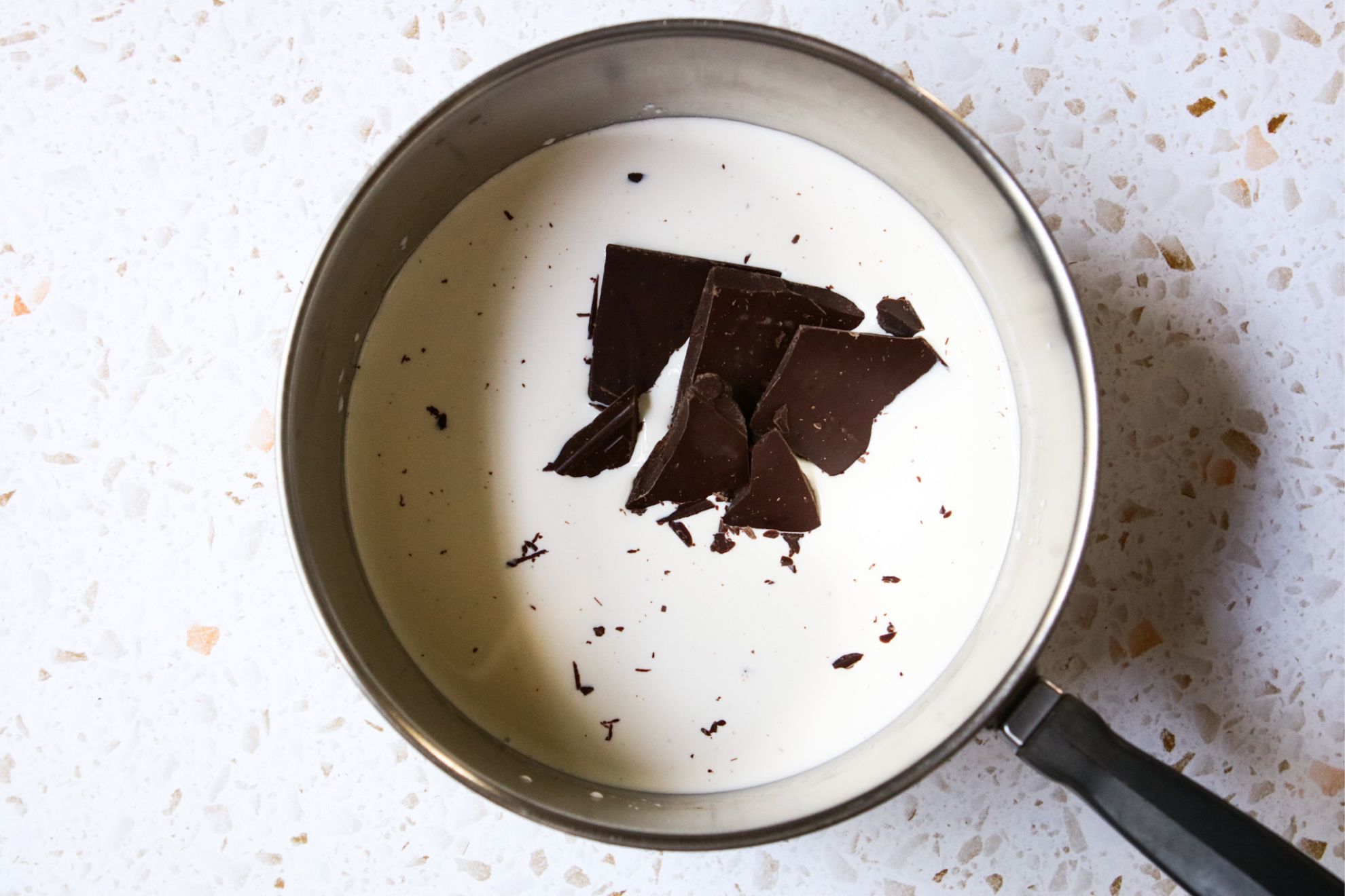 This is an overhead horizontal image of a stainless steel saucepan with milk and chocolate bar pieces in it. The pan sits on a white and tan terrazzo surface.