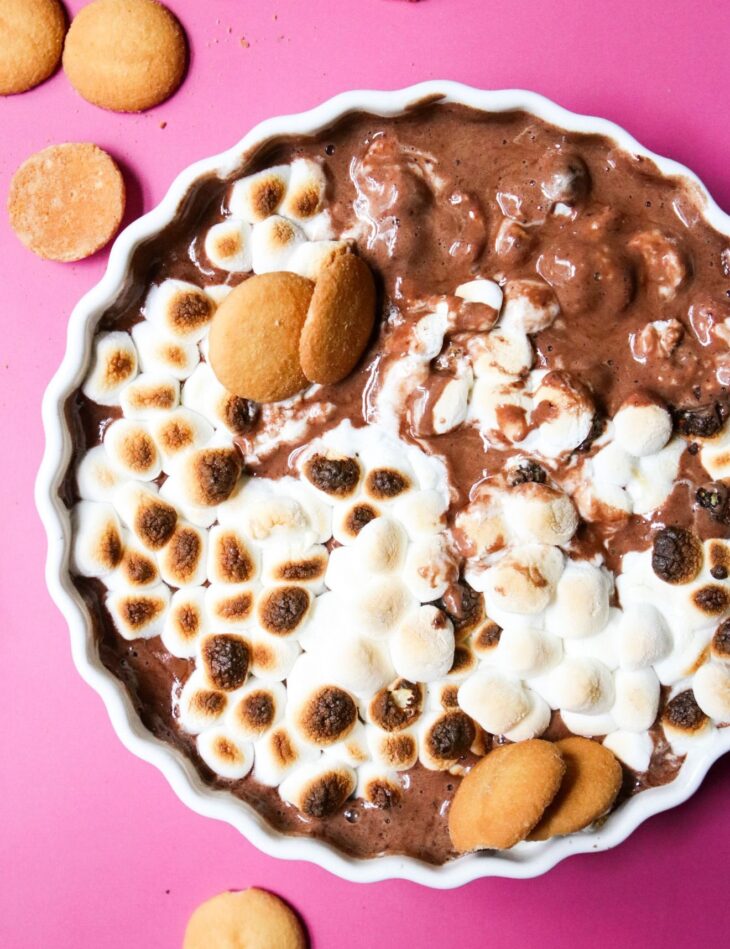 This is an overhead vertical image of a white ceramic scalloped pie dish on a deep pink surface. In the pie dish is a melty, gooey chocolate topped with toasted marshmallows. Nilla wafers are scattered on the pink surface and a few are dipped into the chocolate dip.