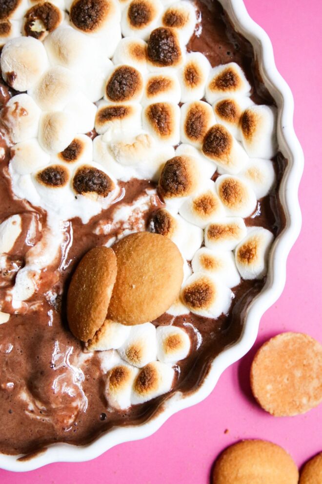 This is an overhead vertical image of a white ceramic scalloped pie dish on a deep pink surface. In the pie dish is a melty, gooey chocolate dip topped with toasted marshmallows. The image is a close up view of the hot chocolate dip. Nilla wafers are scattered on the pink surface and a couple are dipped into the chocolate dip.
