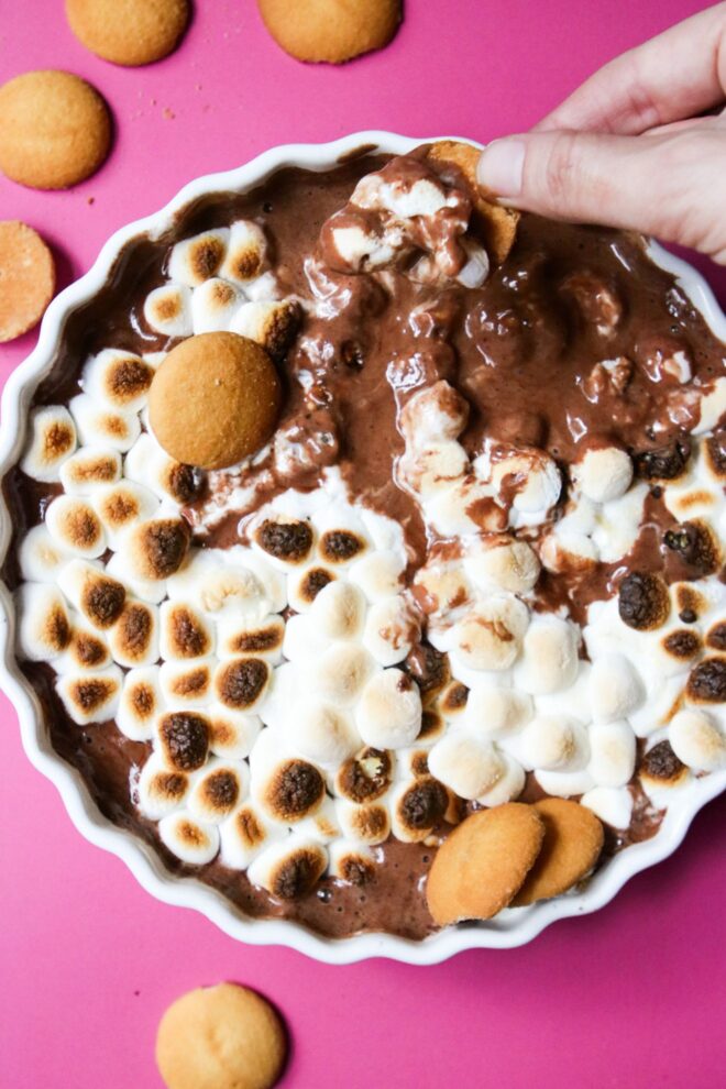 This is an overhead vertical image of a white ceramic scalloped pie dish on a deep pink surface. In the pie dish is a melty, gooey chocolate topped with toasted marshmallows. Nilla wafers are scattered on the pink surface and a few are dipped into the chocolate dip. A hand is coming in from the right top corner of the image and is holding a Nilla wafer, dipping it into the chocolate.