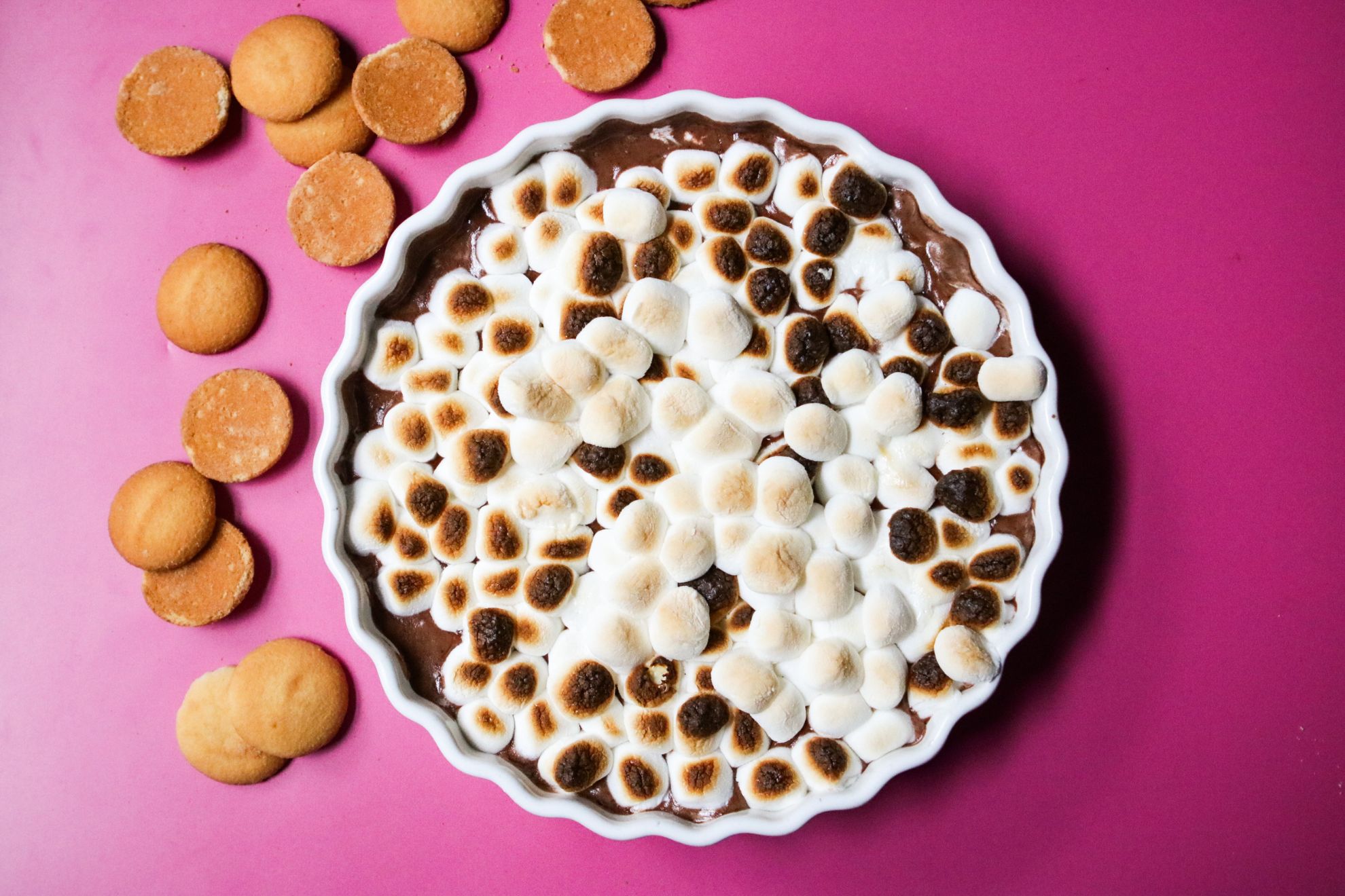 This is an overhead horizontal image of a white ceramic scalloped pie dish sitting on a deep pink surface. In the pie dish is a chocolate mousse spread out in an even layer across the bottom of the dish and topped with toasted mini marshmallows. Nilla wafer cookies are on the pink surface to the left of the pie dish.