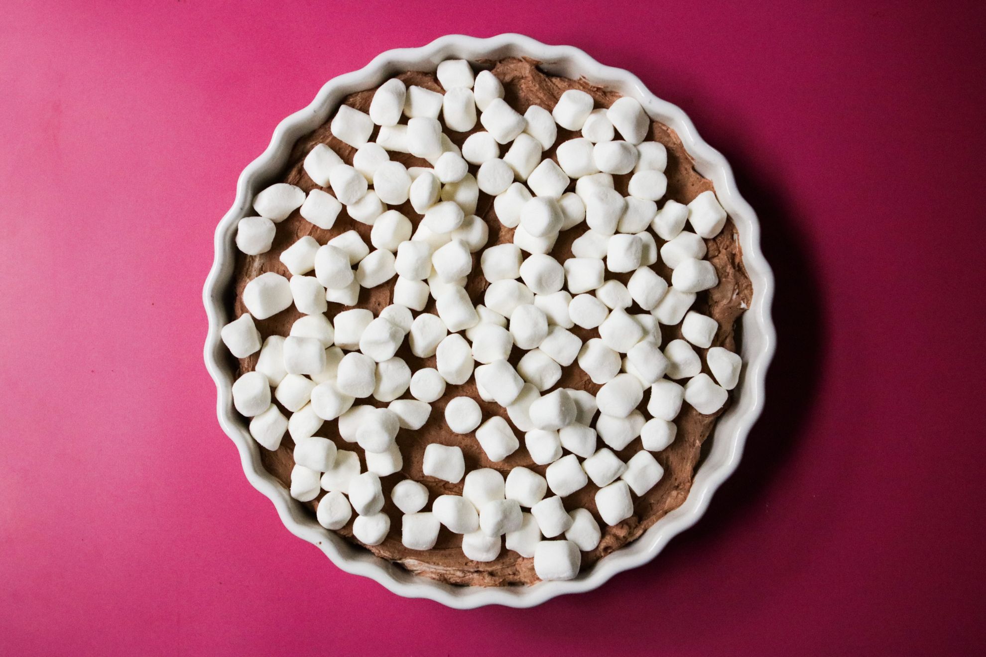This is an overhead horizontal image of a white ceramic scalloped pie dish sitting on a deep pink surface. In the pie dish is a chocolate mousse spread out in an even layer across the bottom of the dish and topped with mini marshmallows.