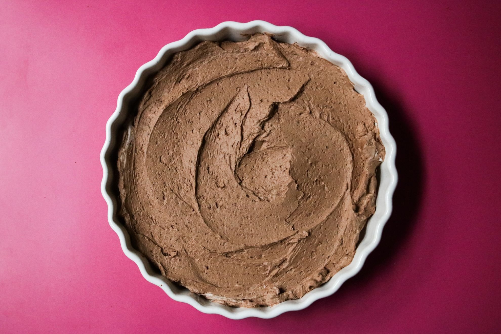 This is an overhead horizontal image of a white ceramic scalloped pie dish sitting on a deep pink surface. In the pie dish is a chocolate mousse spread out in an even layer across the bottom of the dish.