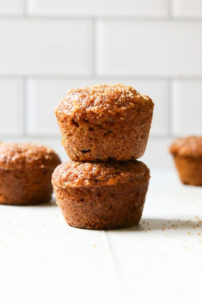 This is a vertical image looking at a stack of two mini muffins from the side. The stack sits on a white surface with more mini muffins blurred in the background. The top of the mini muffin has a brown sugar coating. In the far background are blurred white subway tiles.