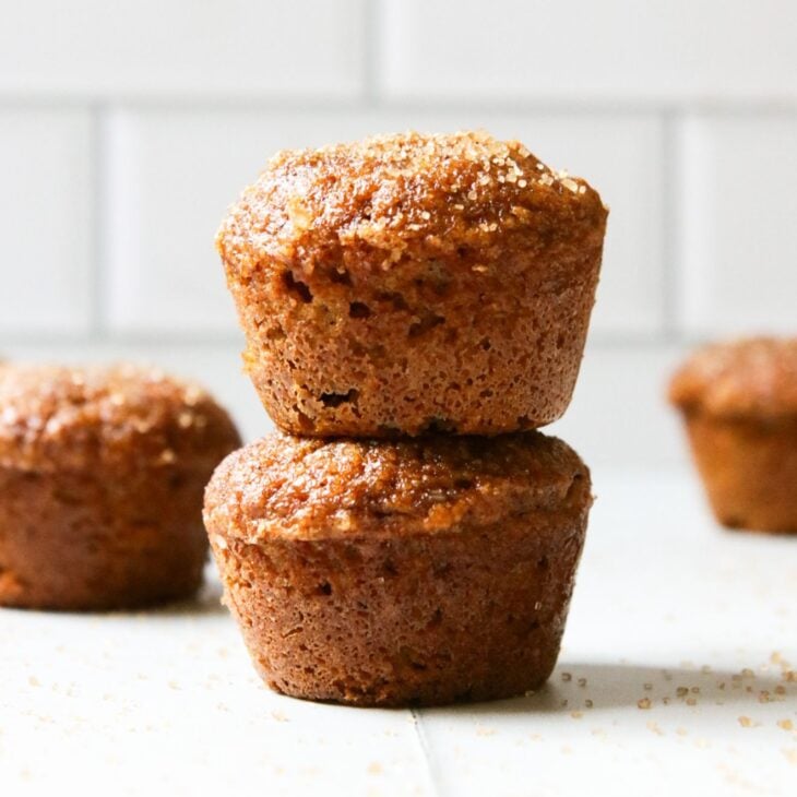 This is a vertical image looking at a stack of two mini muffins from the side. The stack sits on a white surface with more mini muffins blurred in the background. The top of the mini muffin has a brown sugar coating. In the far background are blurred white subway tiles.