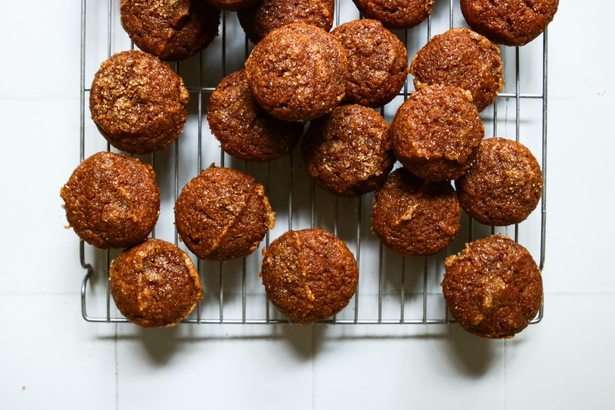 This is an overhead horizontal image of mini pumpkin muffins on a silver cooling rack. The muffins are a rich pumpkin-y color and topped with a brown sugar. Some muffins are stacked on top of others. The cooling rack sits on a white square tile surface.