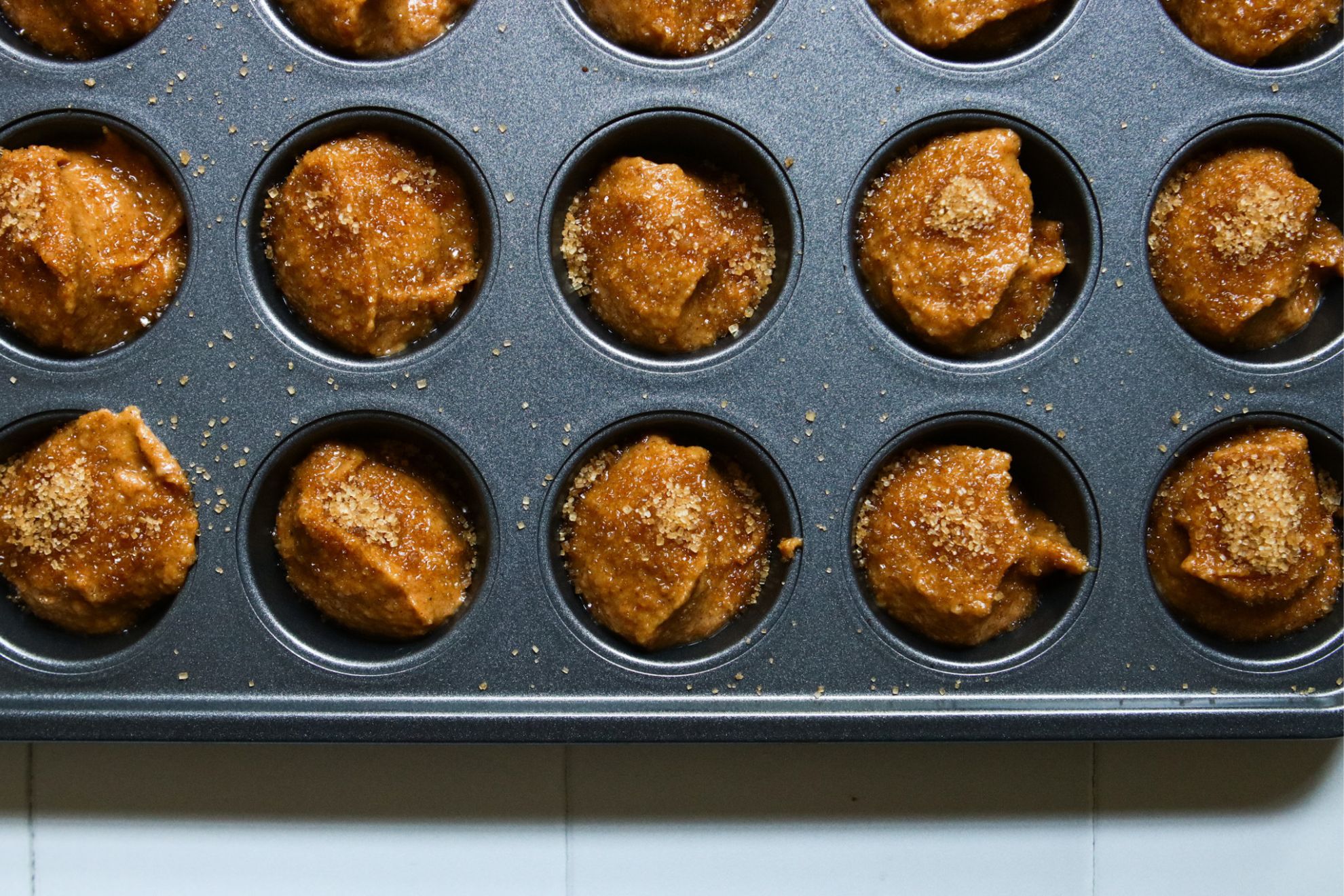 This is an overhead horizontal image focusing on the bottom two rows of a 24 cup mini muffin tin. The tin cups are filled with raw pumpkin muffin batter sprinkled with a light brown sugar. The tin sits on a white square tile surface.