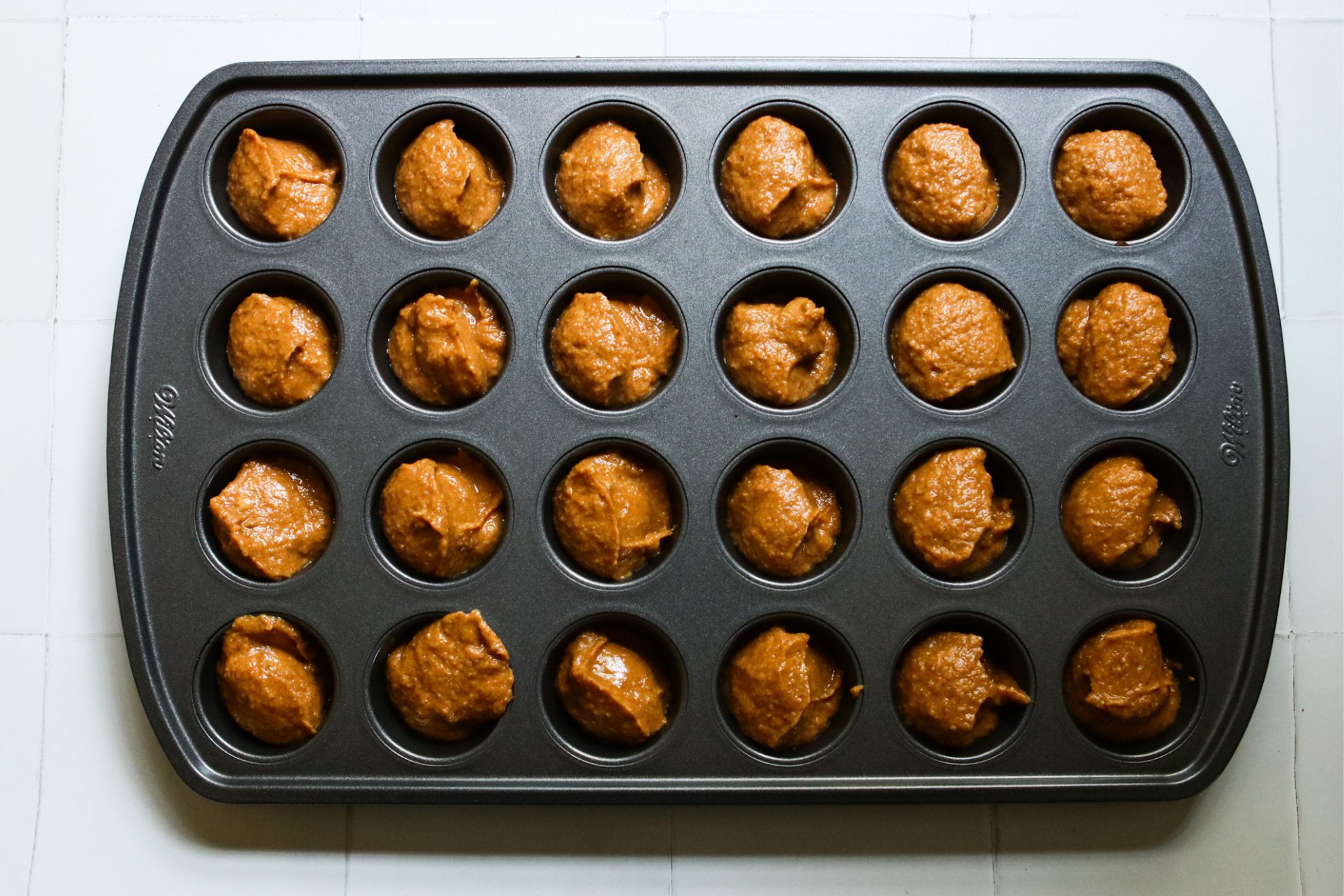 This is an overhead horizontal image of a 24 cup mini muffin tin. The tin cups are filled with raw pumpkin muffin batter. The tin sits on a white square tile surface.