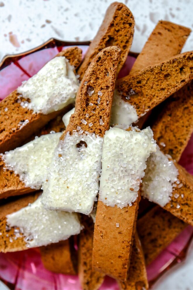 This is an overhead vertical image of orange-brown colored biscotti dipped in white chocolate and sprinkles with clear/white sprinkles. The biscotti are stacked on a clear deep pink plate on a white terrazzo surface.
