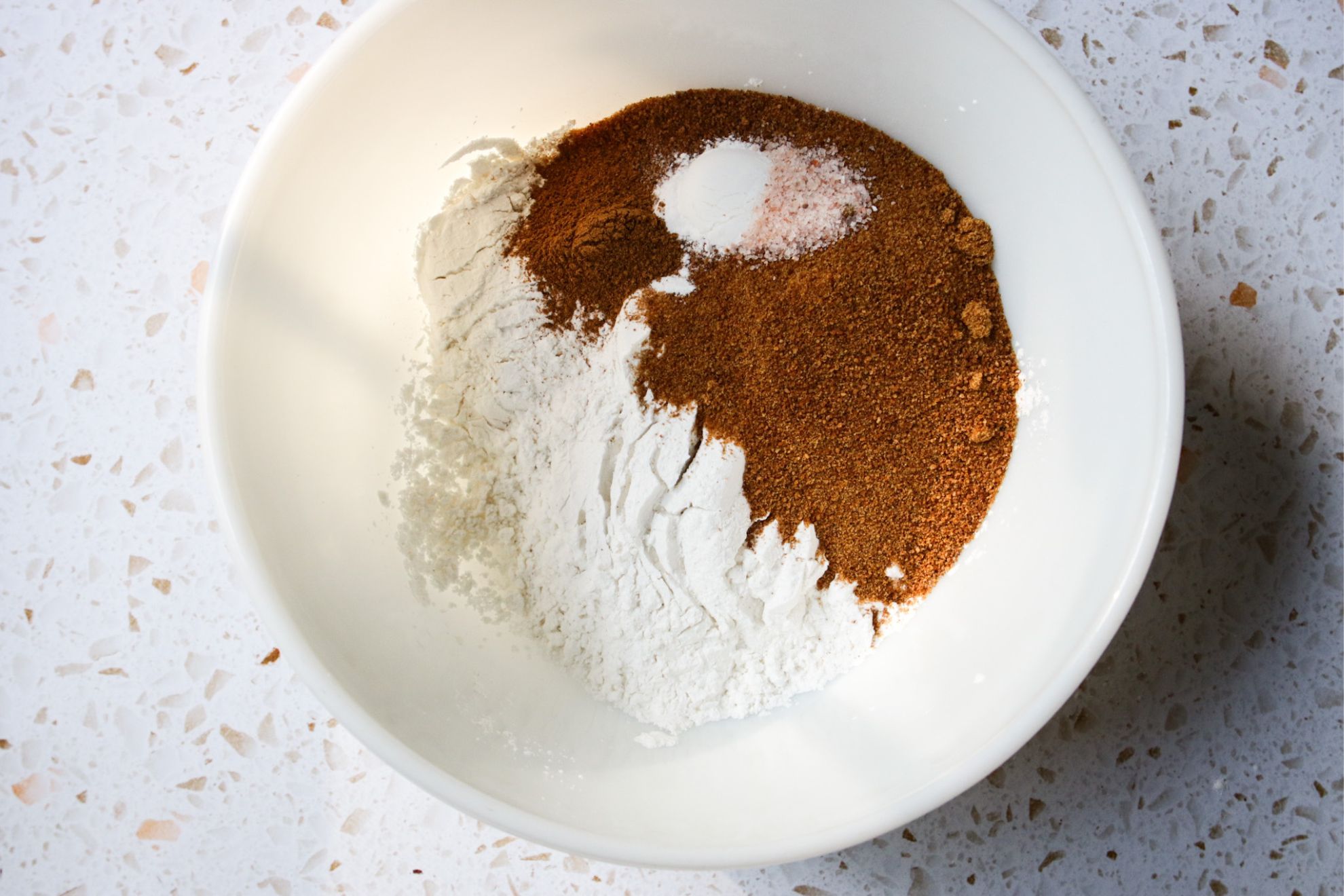 This is a horizontal overhead image of a large white glass bowl on a white and tan terrazzo surface. In the bowl is white flour, coconut sugar, baking powder, pumpkin spice and salt.