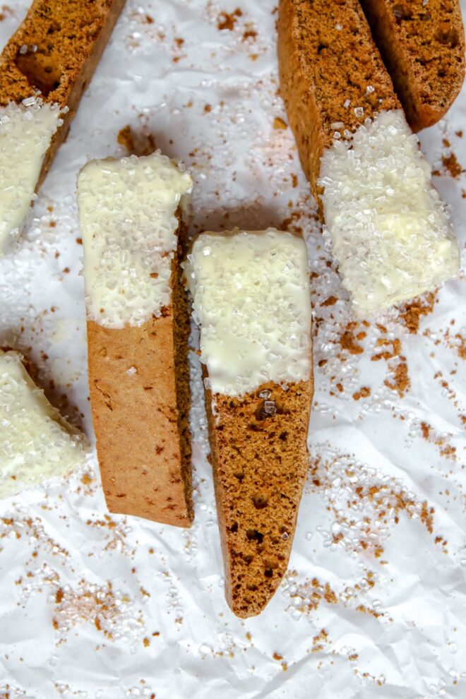 This is an overhead vertical image of orange-brown colored biscotti dipped in white chocolate and sprinkled with clear/white sugar sprinkles. The biscotti sits on a white piece of parchment paper with crumbs and more sprinkles around it.