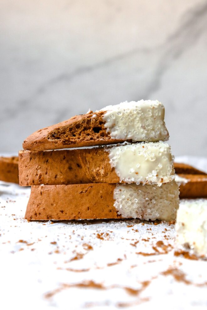 This is a vertical image looking at a stack of three pumpkin biscotti from the side. The stack sits on a white parchment paper with lots of crumbs on it. The biscotti is dipped in white chocolate and has white sugar sprinkles on it. More biscotti is blurred in the background against a marble backdrop.