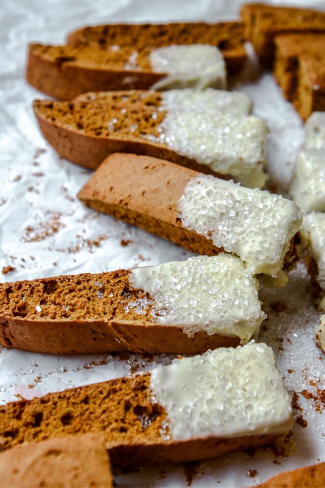 This is a vertical image looking at a row of pumpkin biscotti from the side. The biscotti has dipped white chocolate and sprinkled with clear/white sprinkles. The biscotti sit on a white parchmnet paper.