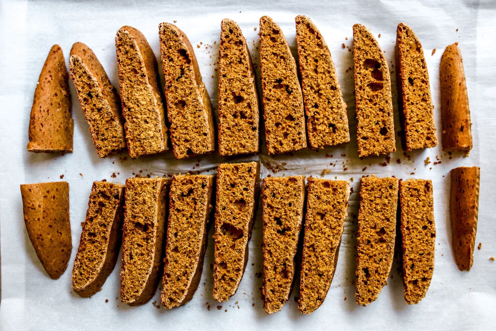 This is an overhead vertical image of a log of biscotti cut into 20 pieces and leaning on their sides. The biscotti cookies lay on a white piece of parchment paper.