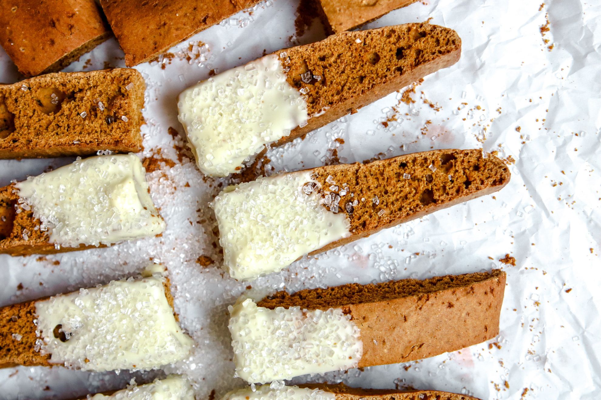 This is an overhead horizontal image of an orange-brown colored biscotti on a white piece of parchment paper. The biscotti is dipped in white chocolate and sprinkled with a clear/white sugar sprinkles.