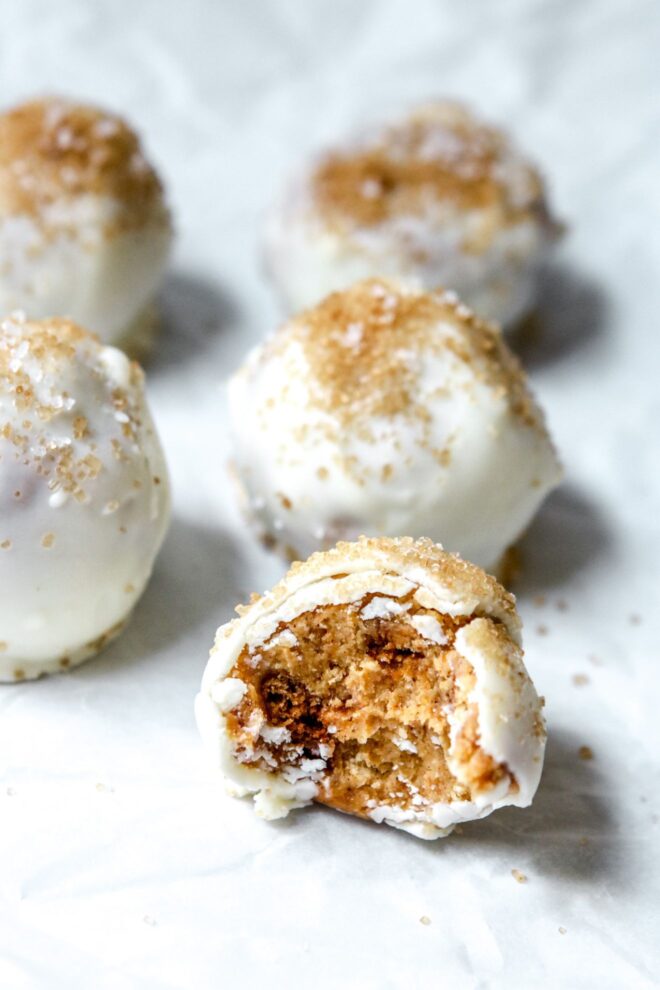 This is a vertical image looking at a ball coating in white chocolate and sprinkled with a tan sugar. The ball has a bite taken out of it and sits on a white piece of parchment paper with more balls behind it.