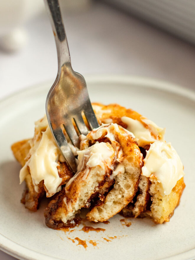 This is a vertical image looking at a cinnamon roll from the side with shite vanilla frosting on it. A fork is coming in from the top of the image and piercing the center of the roll. The cinnamon roll sits on a small white plate on a white marble counter with a white casserole dish blurred in the background.