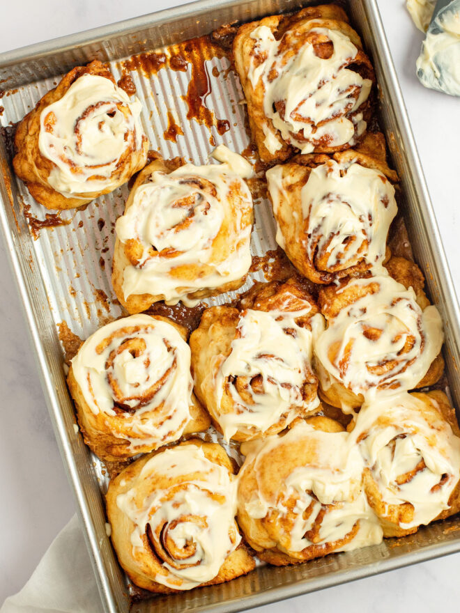 This is an overhead vertical image of a 9x13 rectangle pan on a white marble surface. The pan is vertical and at an angle tilting to the left side of the image. In the pan are cinnamon rolls with creamy white frosting on them. A grey rubber spatula with frosting on it is coming into the image from the top right corner.