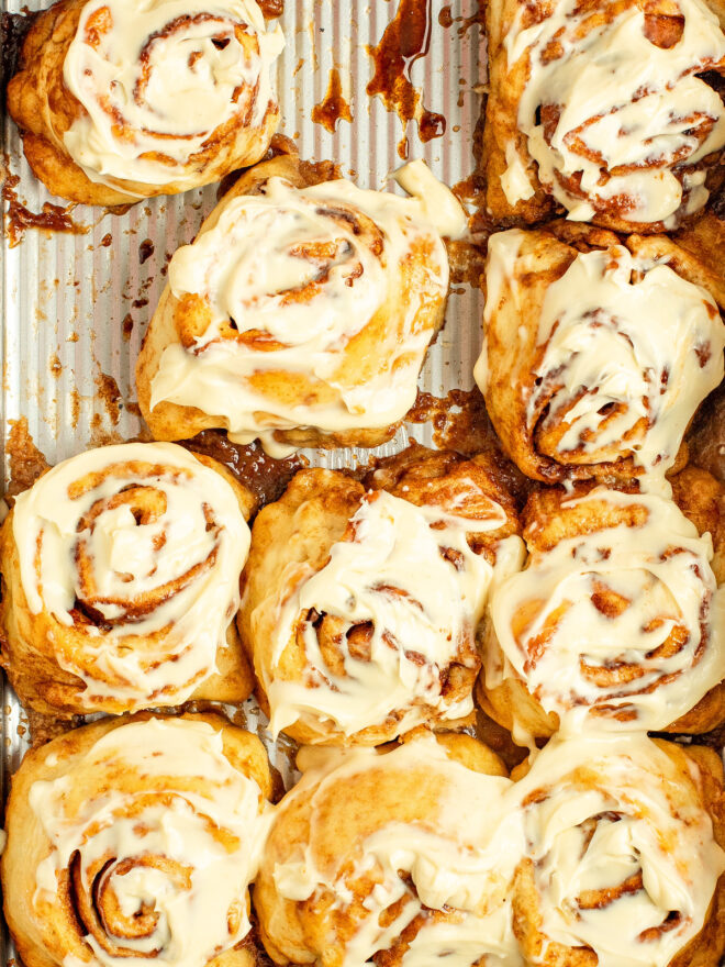 This is an overhead vertical image inside a 9x13 rectangle pan with cinnamon rolls topped with creamy white frosting. A couple cinnamon rolls are missing displaying the gooey cinnamon drips that have fallen from the cinnamon rolls.