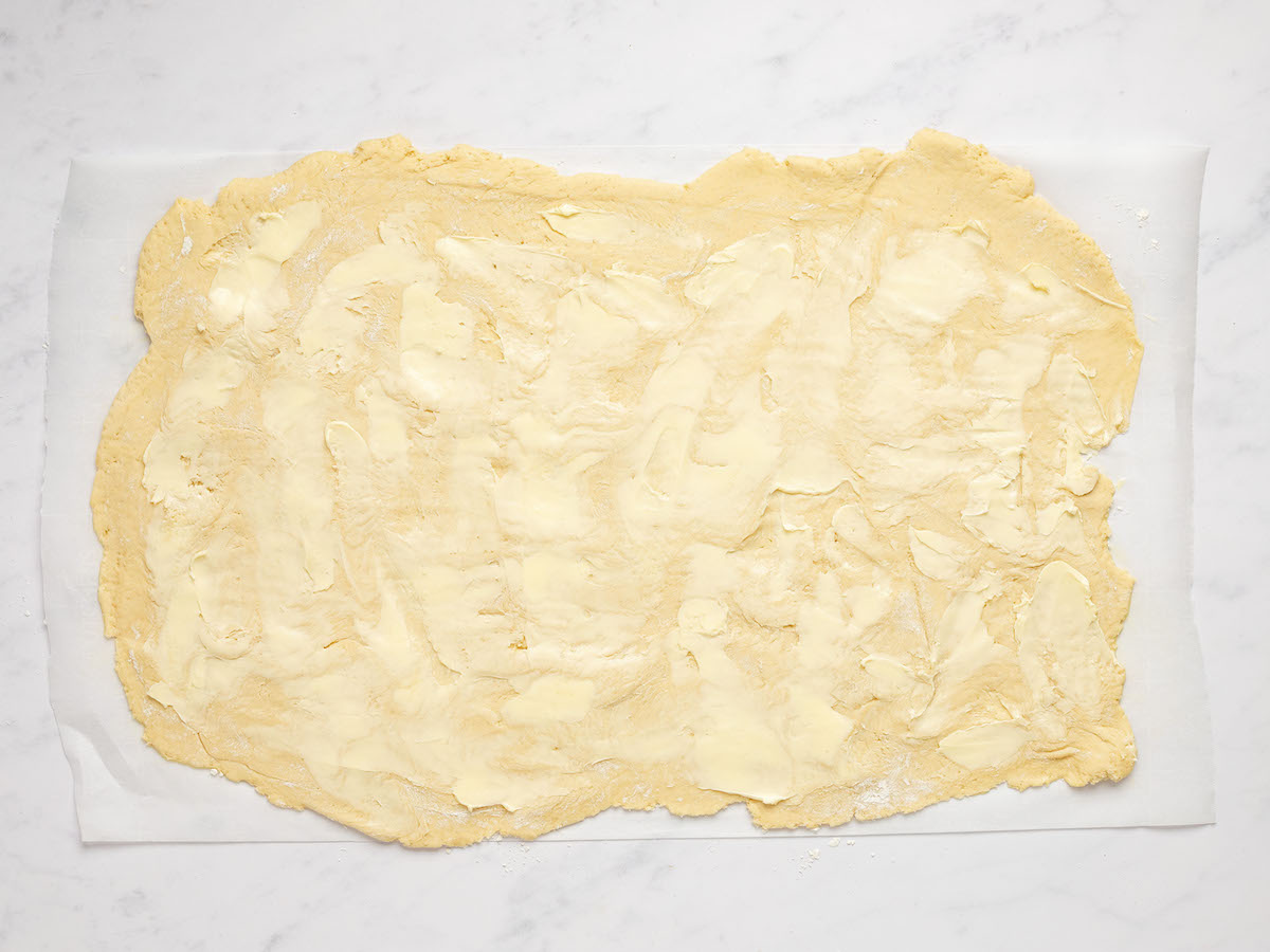 This is an overhead horizontal image of dough rolled out in a horizontal rectangle. The dough lies on a white piece of parchment paper on a white marble counter. The dough has softened butter spread across it.