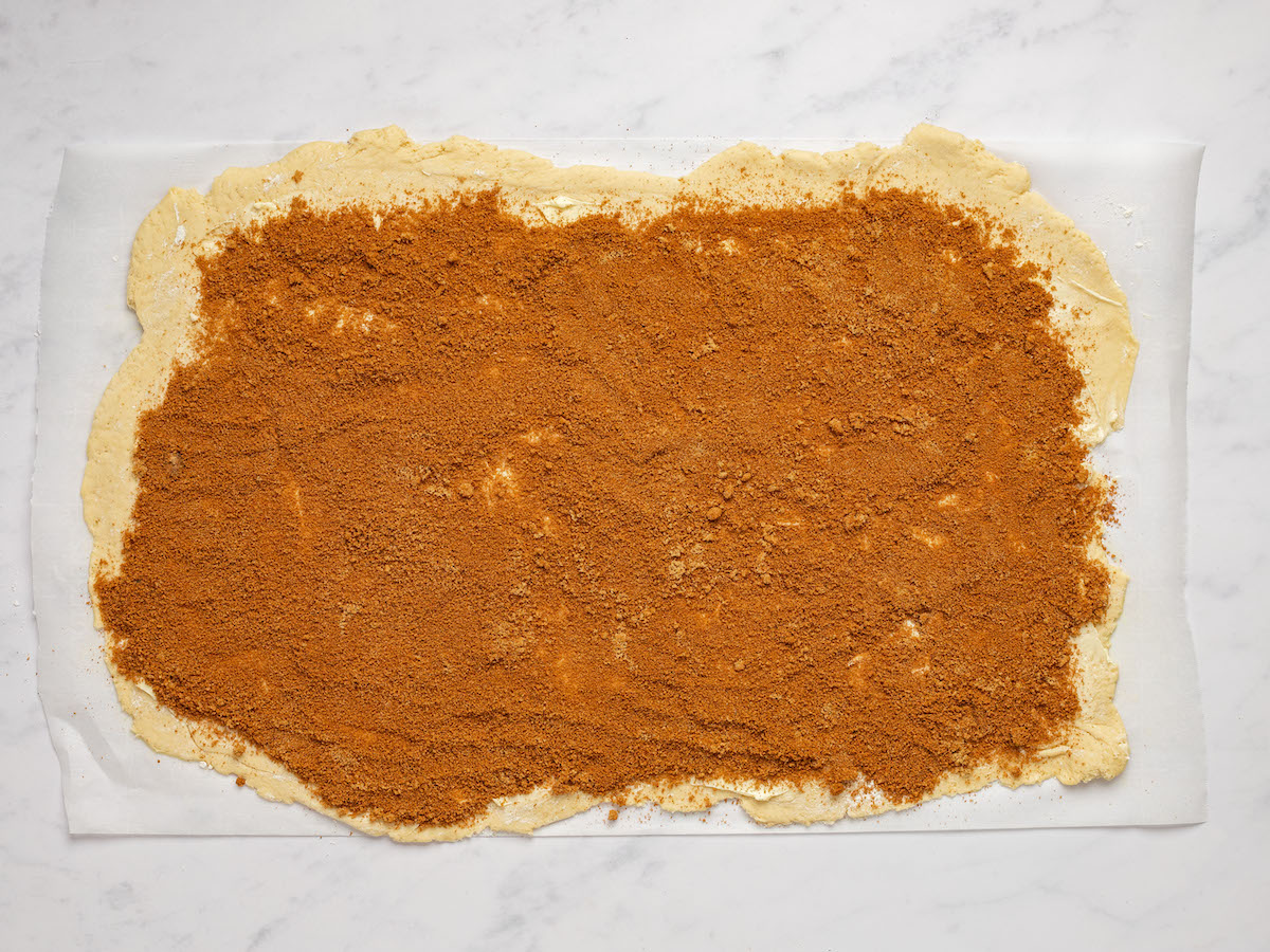 This is an overhead horizontal image of dough rolled out in a horizontal rectangle. The dough lies on a white piece of parchment paper on a white marble counter. The dough has cinnamon sugar sprinkled across it.
