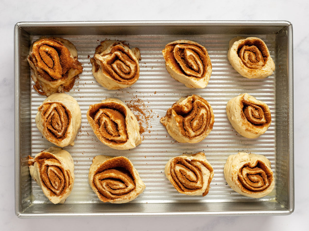 This is an overhead horizontal image of a rectangle pan with 12 cinnamon buns sitting in the pan, 1 - 2 inches apart, with their cinnamon swirl facing up. The pan sits on a white marble surface.