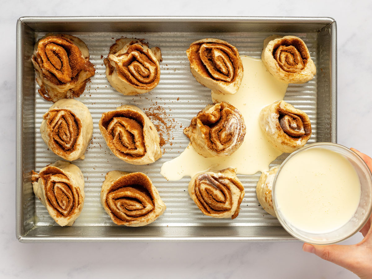 This is an overhead horizontal image of a rectangle pan with 12 raw cinnamon buns sitting in the pan, 1 - 2 inches apart, with their cinnamon swirl facing up. The pan sits on a white marble surface. A hand is coming from the bottom right corner holding a small glass bowl of heavy cream. The cream is slowly pouring into the bottom of the pan around and on top of the cinnamon rolls.