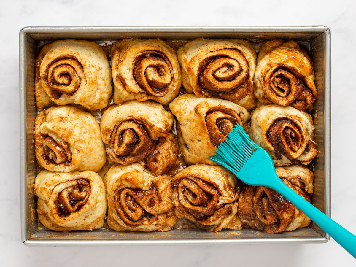 This is an overhead horizontal image of a rectangle pan with 12 baked cinnamon buns sitting in the pan, with their cinnamon swirl facing up. The pan sits on a white marble surface. A blue silicon pastry brush is coming from the bottom right corner holding and brushing the tops of the cinnamon rolls.