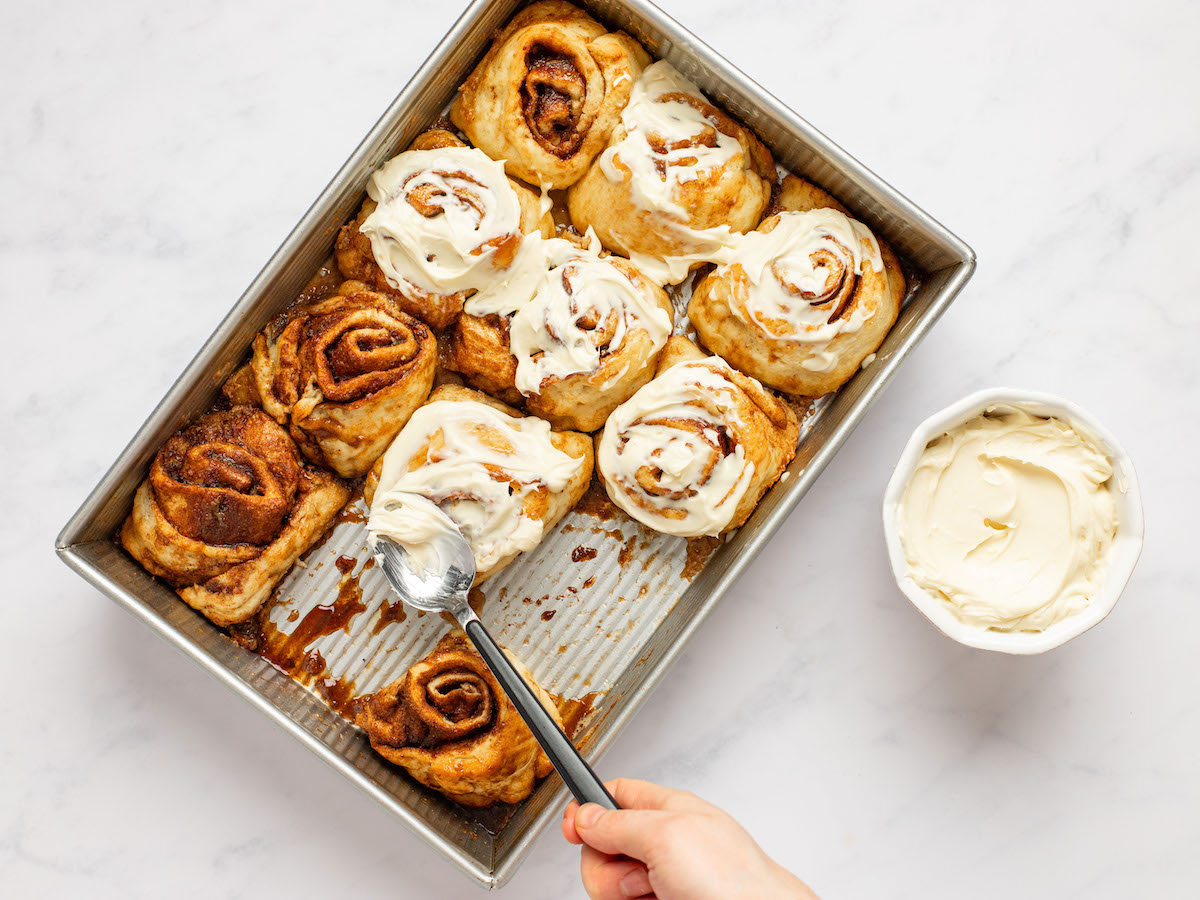 This is an overhead horizontal image of a silver rectangle pan on a white marble surface. The pan is at an angle in the center of the image with a small white bowl of vanilla frosting to the right of the image. Two cinnamon rolls are missing from the pan. A hand is coming from the bottom of the image holding a spoon and spreading the vanilla frosting across the tops of the cinnamon rolls.