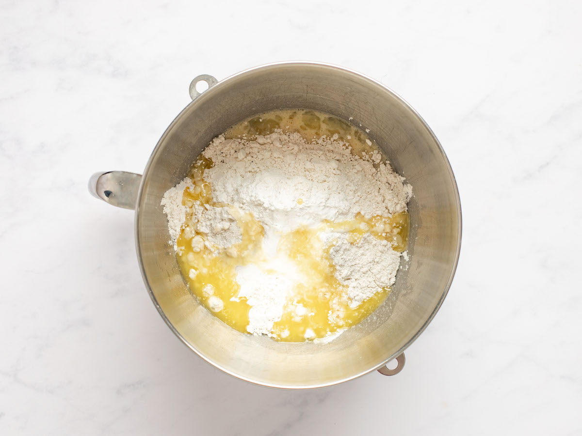 This is an overhead horizontal image of a silver mixing bowl with dry ingredients on top of wet ingredients . The bowl sits on a white marble counter.