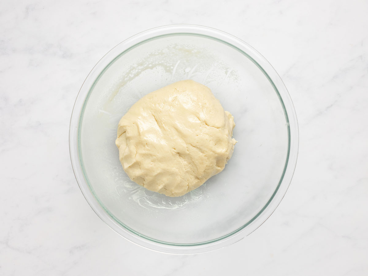 This is an overhead horizontal image of a glass bowl filled with a ball of dough. The bowl sits on a white marble counter.
